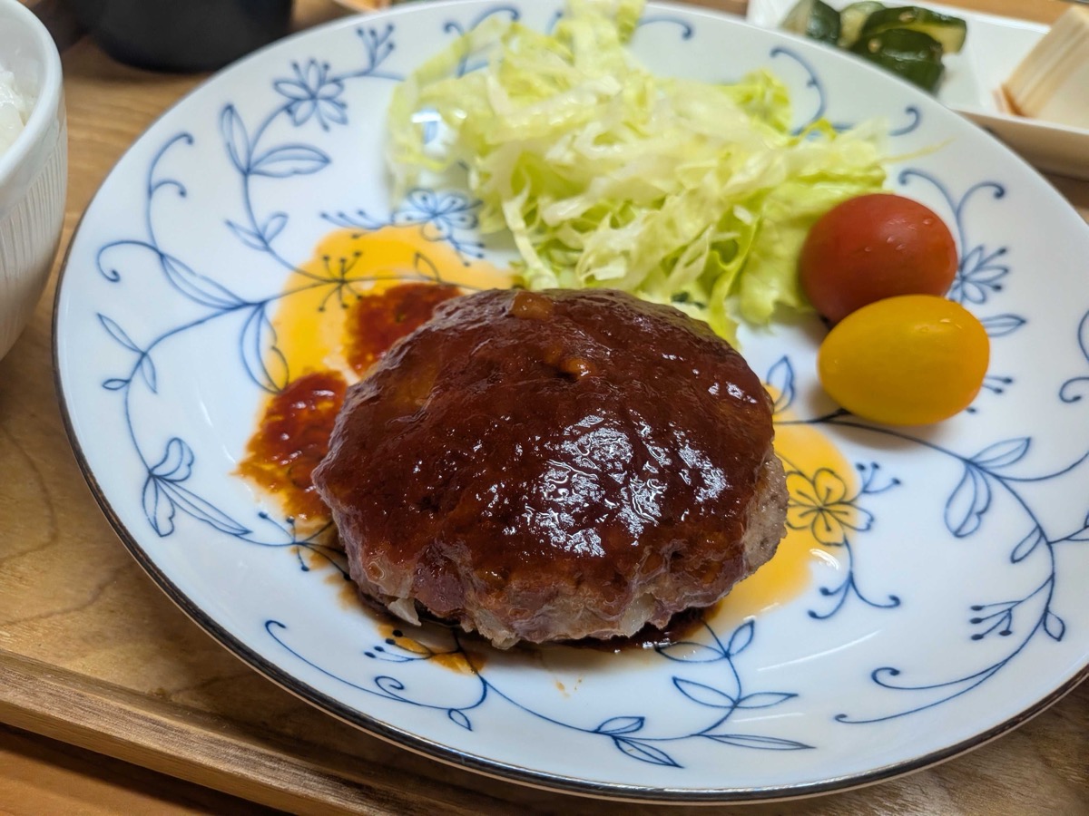 Finished Japanese hamburg steak with glossy sauce, shredded cabbage, and cherry tomatoes on a blue floral plate with rice on the side
