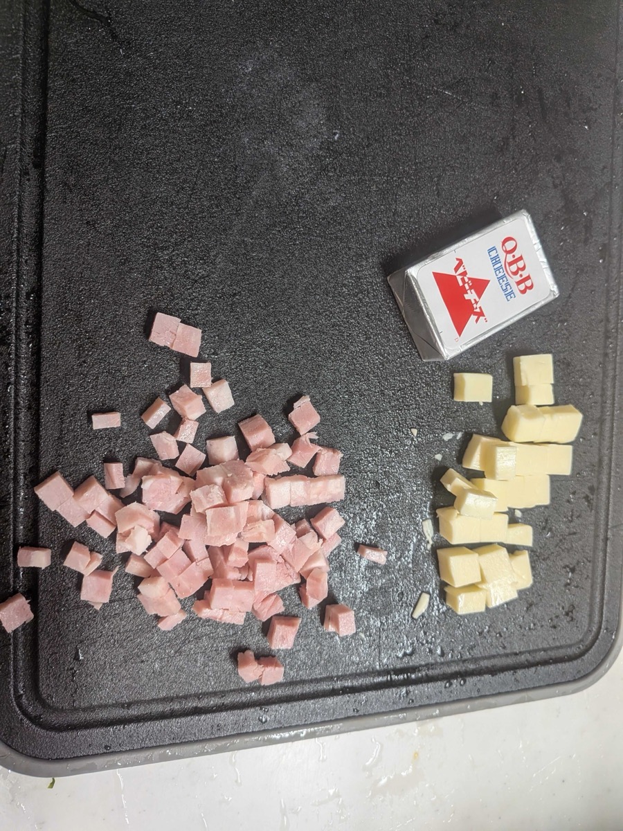 Diced ham and cubed processed cheese on a black cutting board next to a QBB cheese box