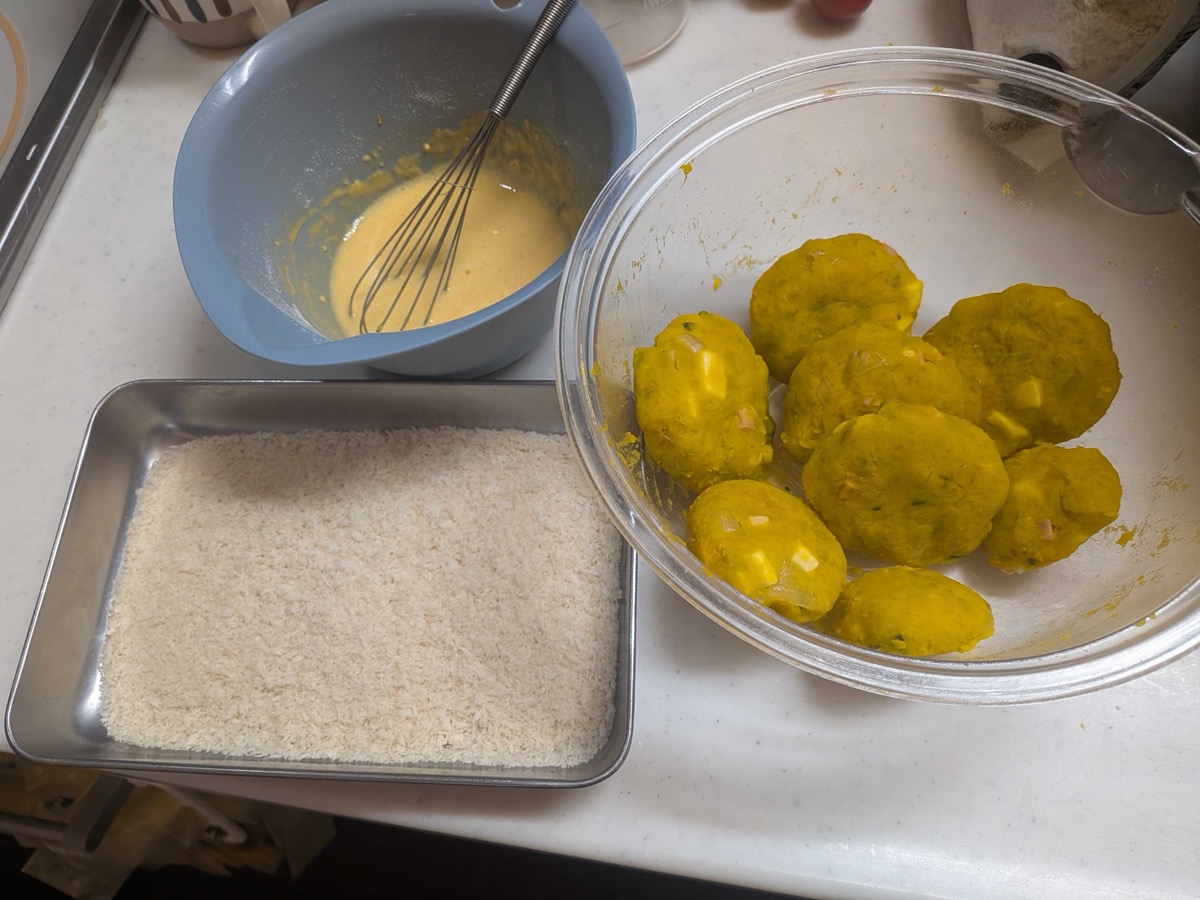 Round shaped kabocha pumpkin croquette patties in a glass bowl next to batter and a tray of panko