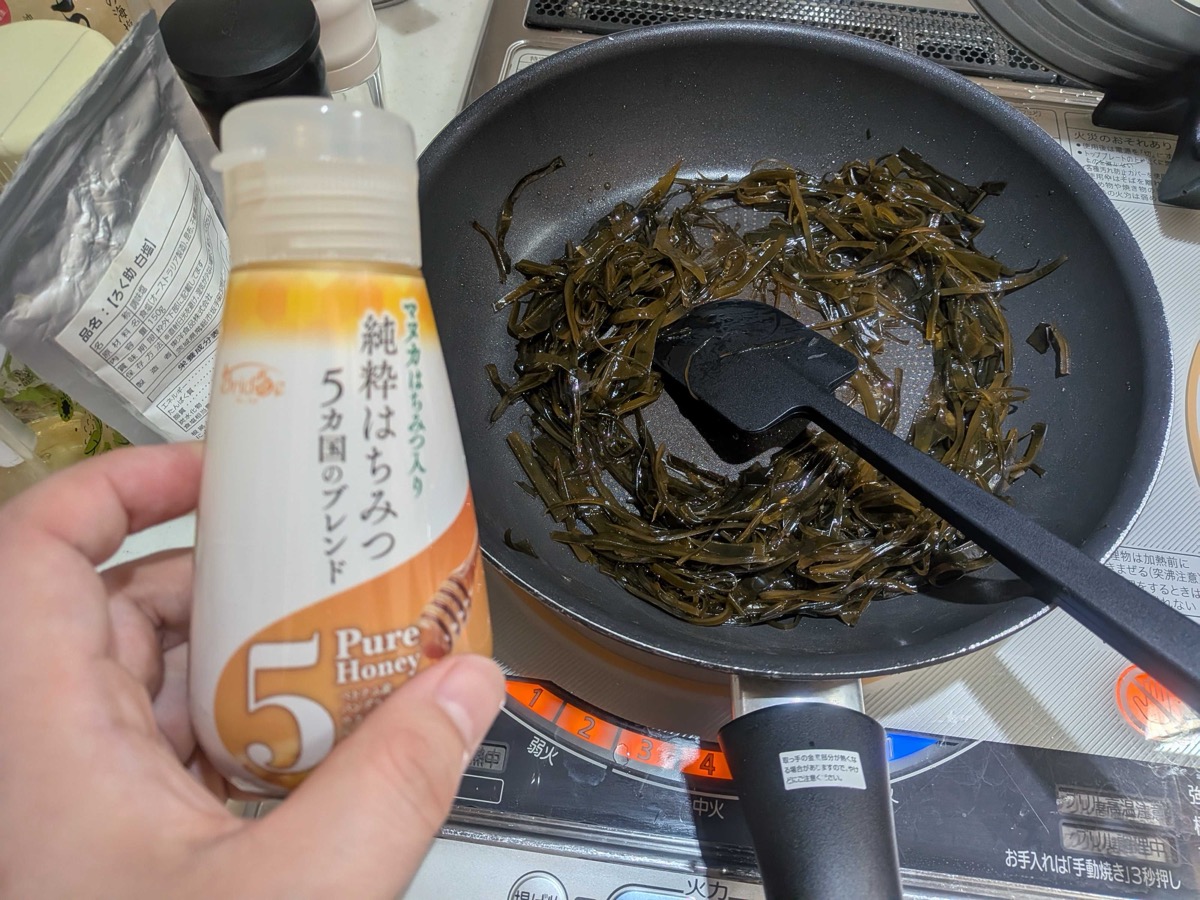 A bottle of Japanese Manuka blend pure honey being held over a pan of soy-simmered kombu strips, ready to be squeezed in to finish the tsukudani glaze