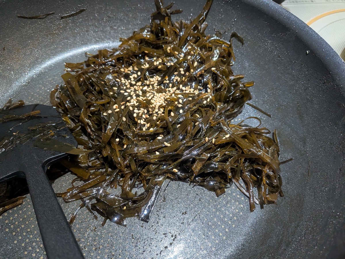 A generous pinch of toasted white sesame seeds sprinkled over the finished glossy kombu no tsukudani in the pan, ready to be stirred through