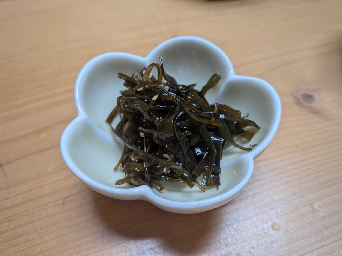 A small white cherry-blossom-shaped Japanese dish holding finished kombu no tsukudani — dark glossy strips of kelp with white sesame seeds scattered through — resting on a light wooden table