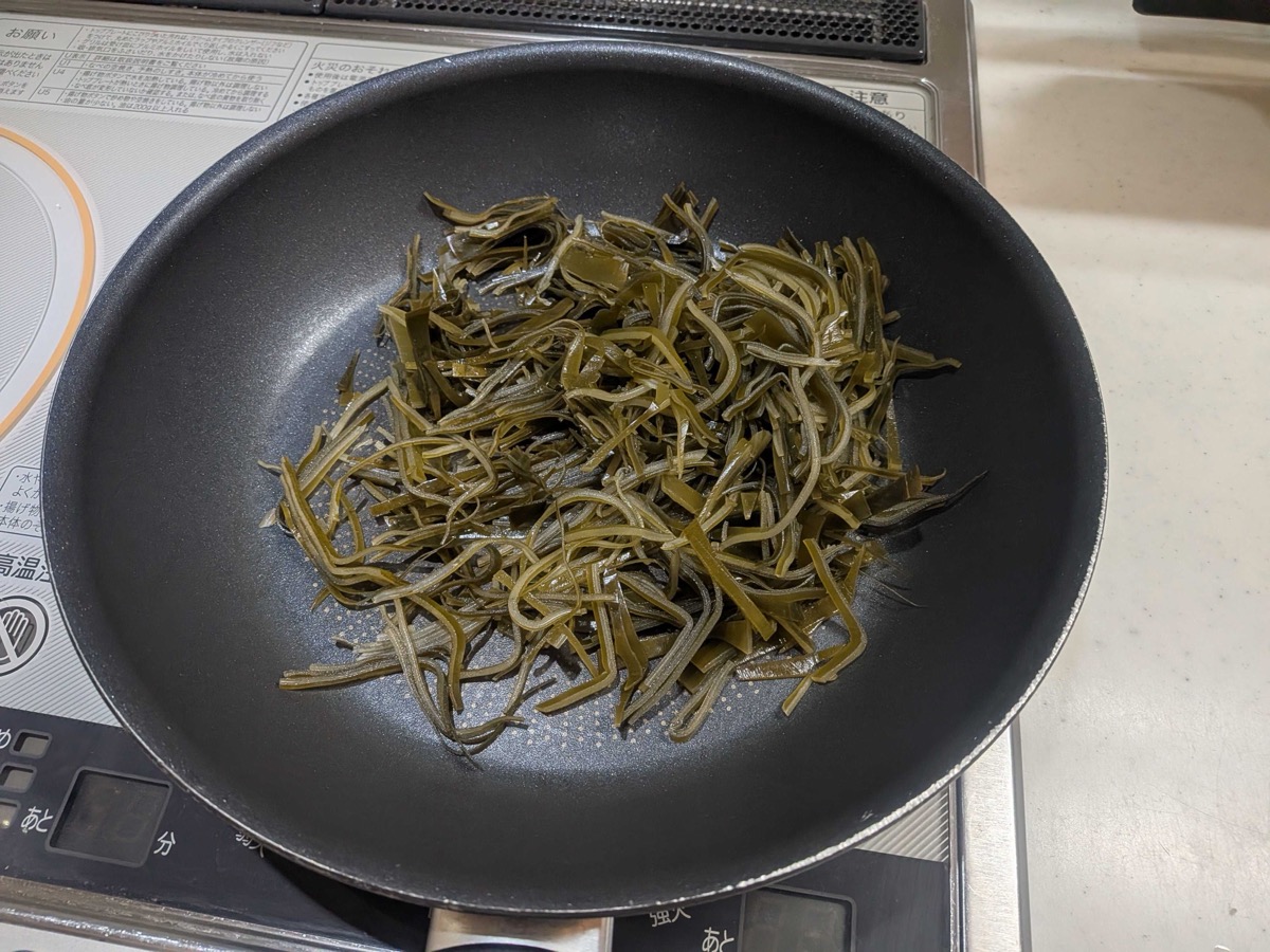 A wide black non-stick frying pan on an IH cooktop holding thinly sliced strips of rehydrated leftover dashi kombu — the starting point for kombu no tsukudani