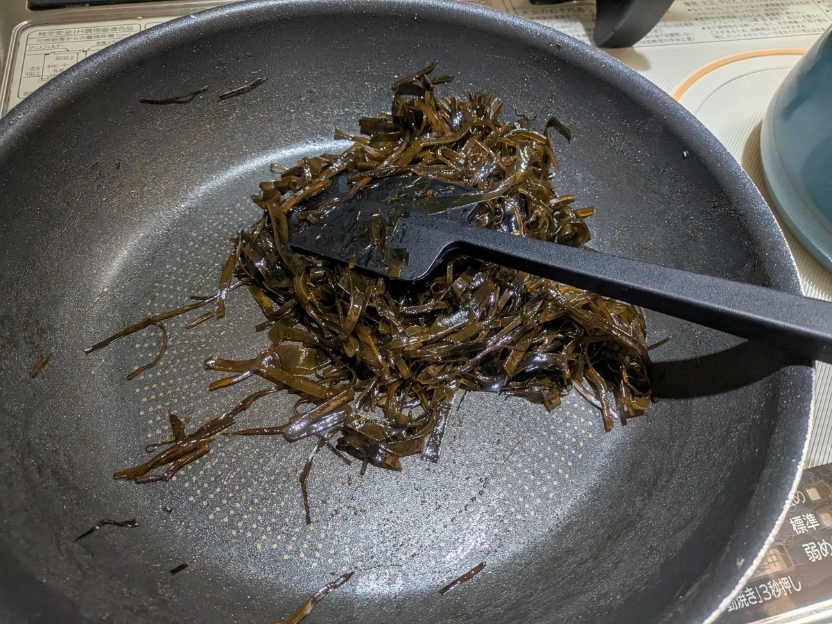 Finished kombu no tsukudani gathered in a mound in the centre of the frying pan — dark, glossy, sticky strips of kelp coated in a rich soy glaze with a black spatula pushing it together