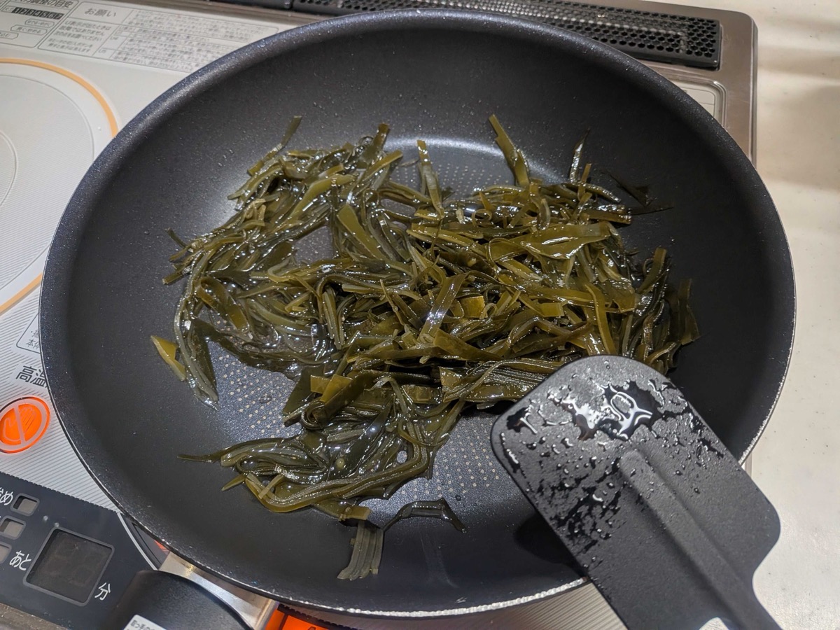 Sliced kombu strips simmering in a shiny mirin-sake-sugar-vinegar liquid in a black frying pan, with a rubber spatula visible on the right — the first covered simmer stage of kombu no tsukudani