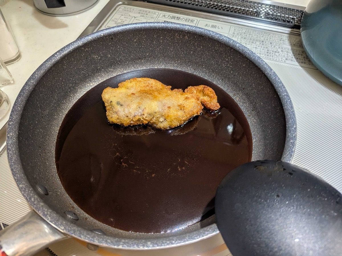 A fried pork cutlet being dipped into the pan of katsudon sauce