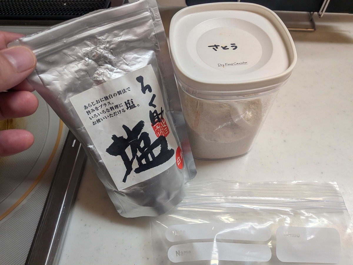 A silver pouch of Japanese Rokusuke shio finishing salt held next to a sugar canister and empty freezer bags on the kitchen counter — the seasoning that gives homemade shio kombu its signature clinging umami finish