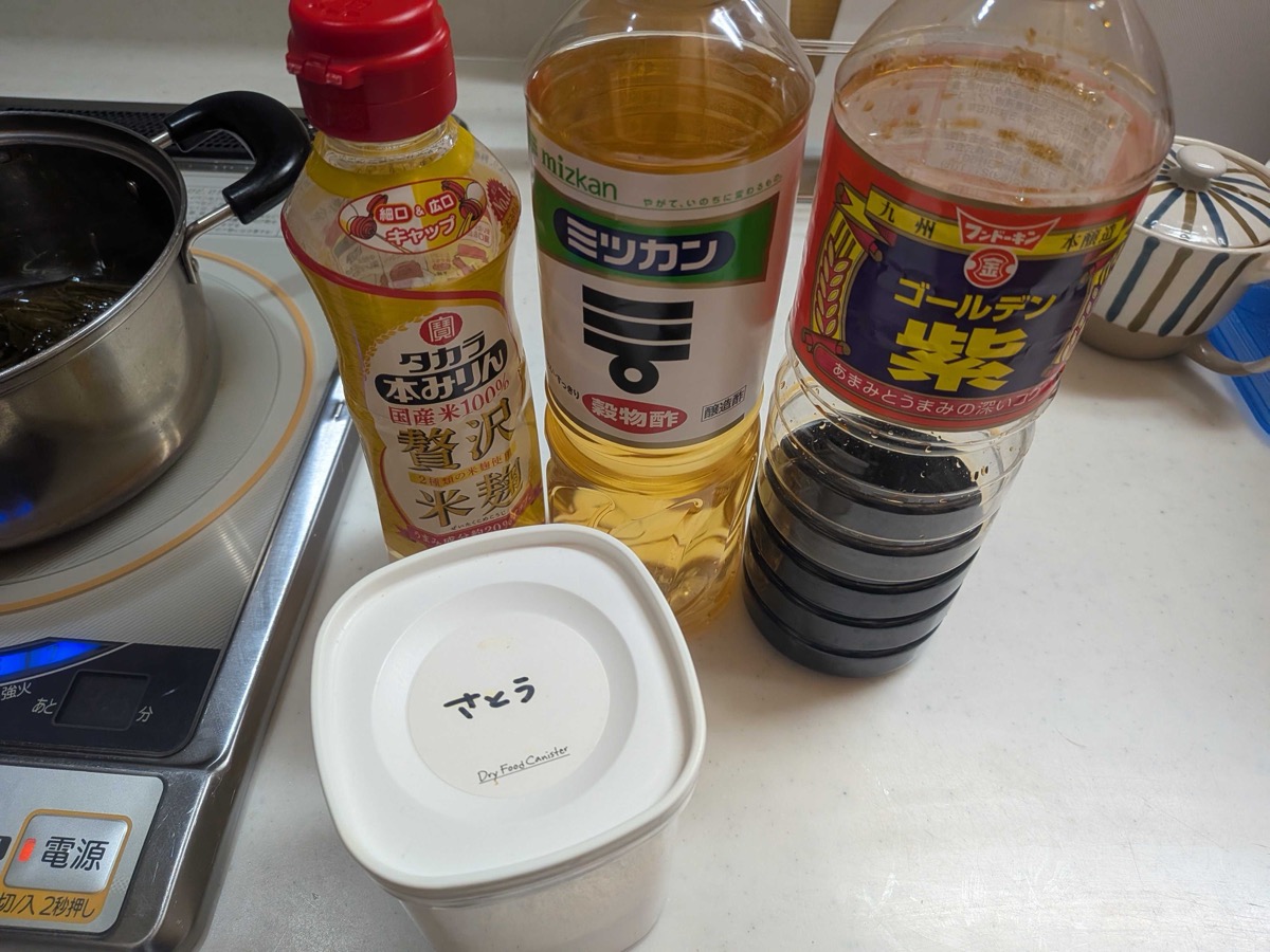 The seasonings for shio kombu lined up on a kitchen counter — a bottle of Takara honmirin, Mizkan rice vinegar, Fundokin Golden Murasaki soy sauce, and a sugar canister — next to a pot with sliced kombu