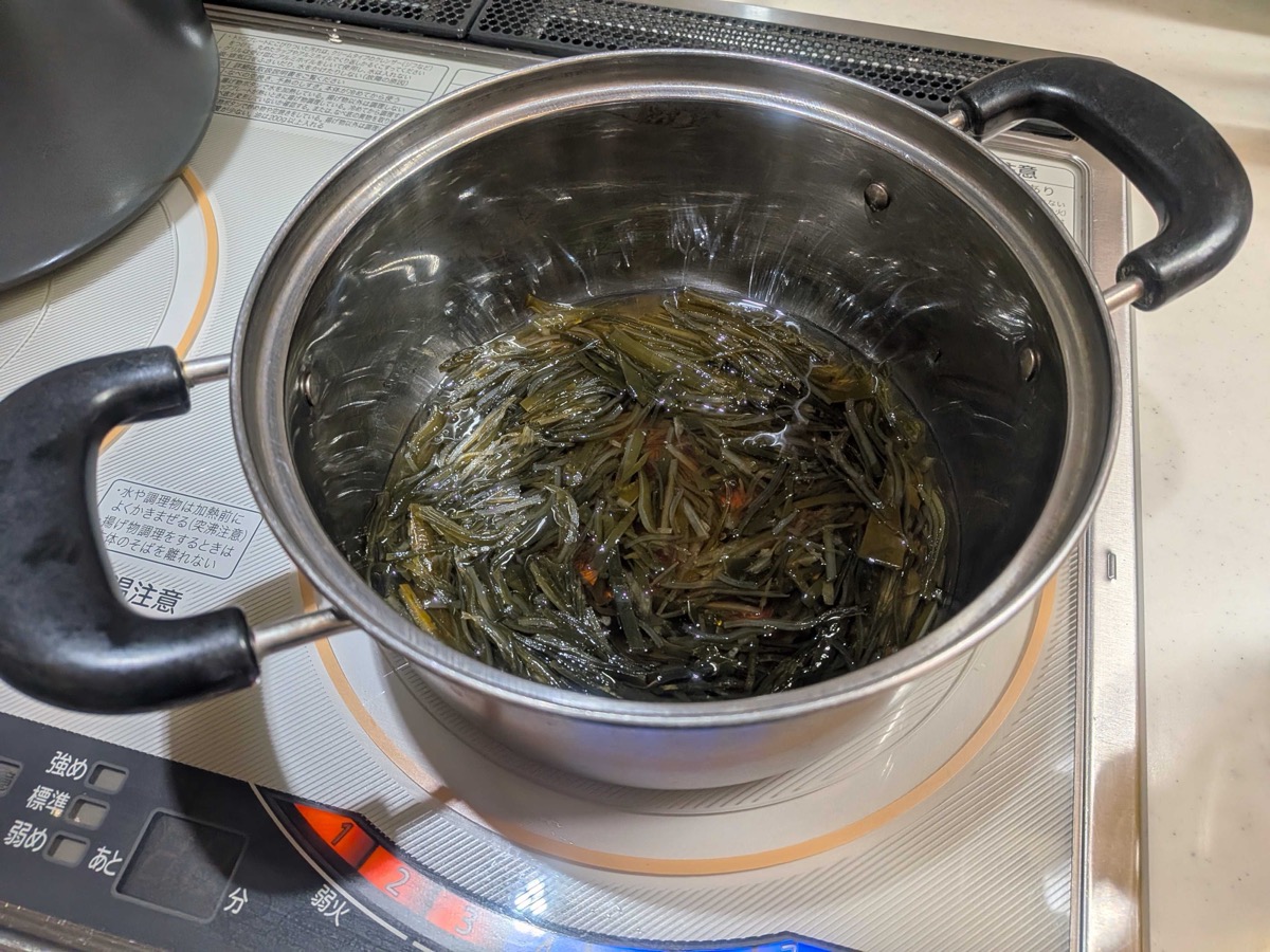 Thin strips of kombu simmering in a stainless steel pot with a dark sweet-soy-mirin liquid on an IH cooktop — the second stage of making Japanese shio kombu