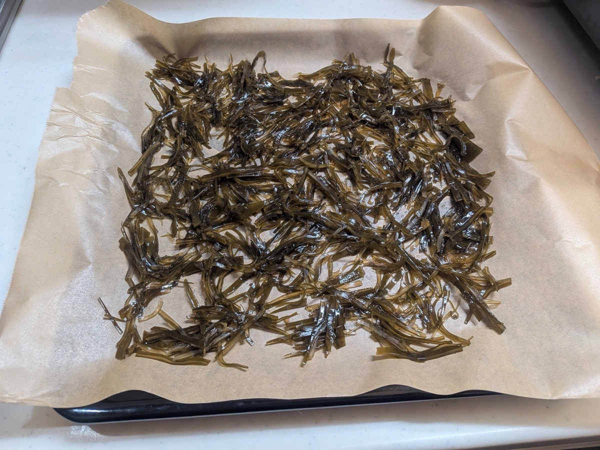 Sticky sweet-soy simmered kombu strips spread in a single loose layer on parchment paper on a black baking sheet, ready to go into a low oven to dry