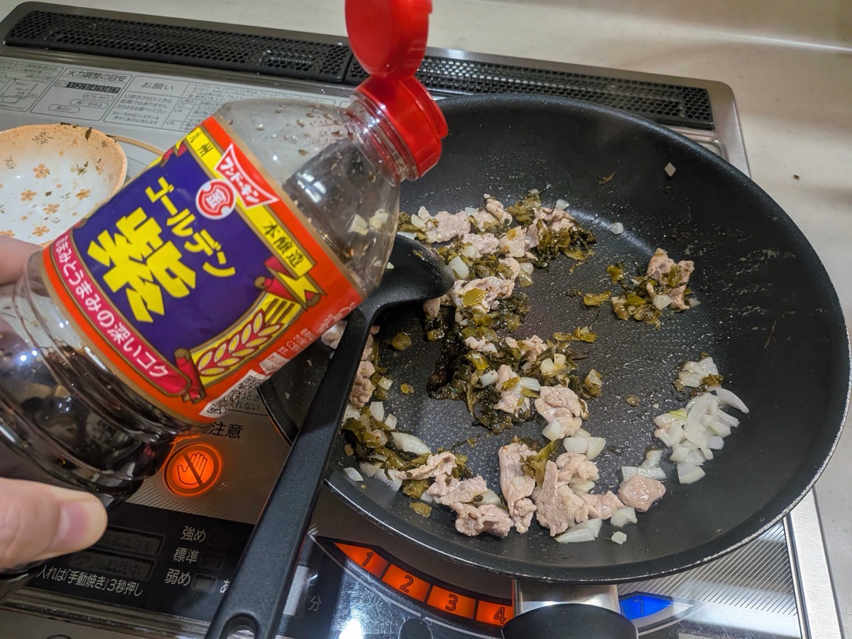 Chopped takana greens and soy sauce being added to the pork and onion in the frying pan