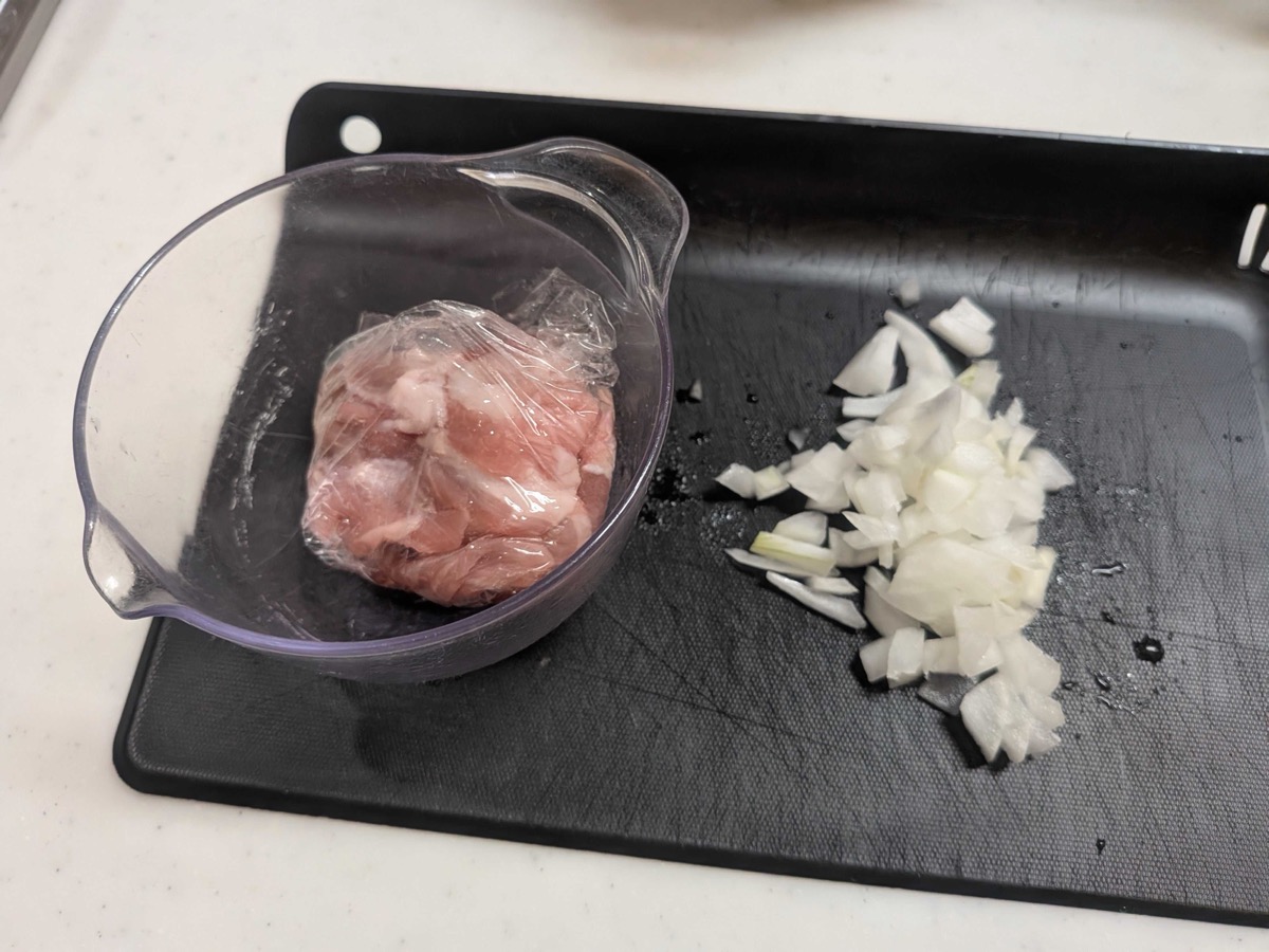 Thinly sliced pork in a bowl next to finely chopped onion on a black cutting board