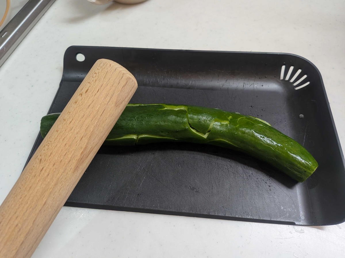A whole Japanese cucumber being smashed on a black cutting board with a wooden rolling pin, cracks visible along the length