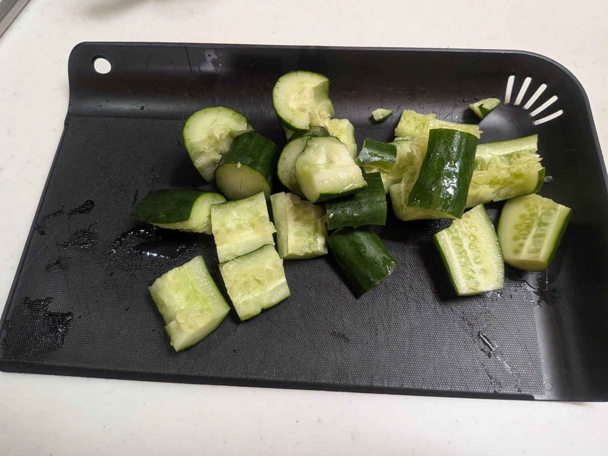 Roughly chopped pieces of smashed cucumber piled on a black cutting board, showing jagged torn edges from being cracked with a rolling pin