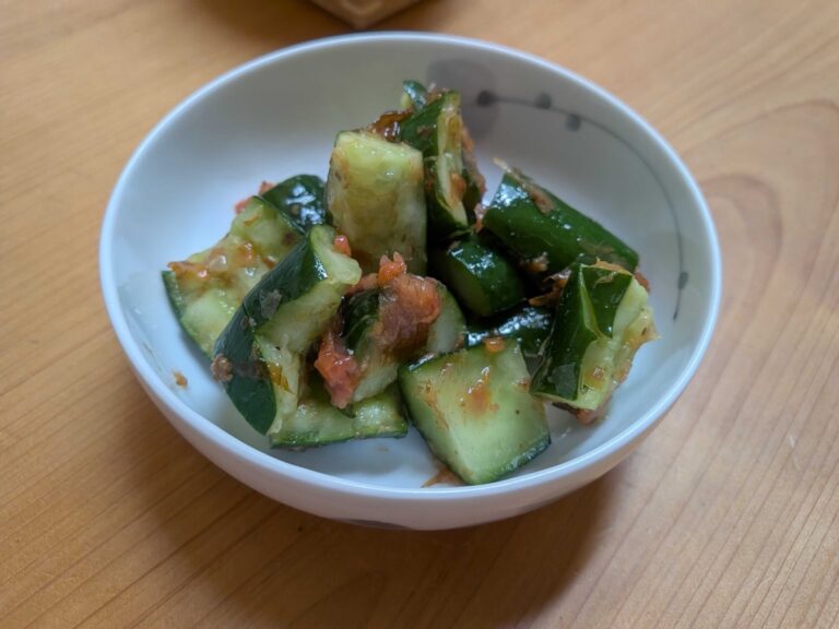 A small white Japanese bowl of finished tataki ume kyuri — smashed cucumber pieces coated in chopped red umeboshi and bonito flakes, sitting on a wooden table