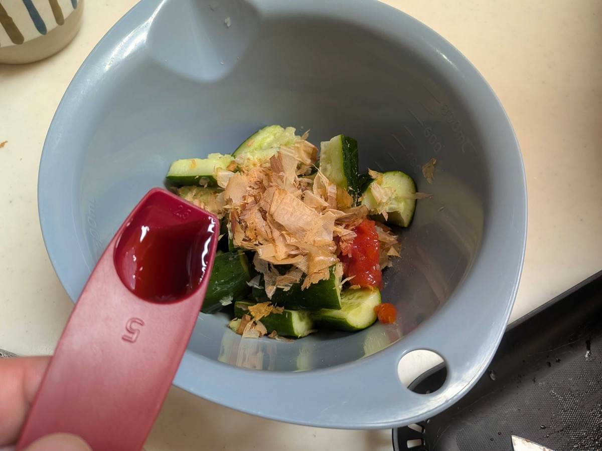 A blue mixing bowl with smashed cucumber, chopped umeboshi, and a mountain of katsuobushi bonito flakes as mentsuyu is added from a red measuring spoon