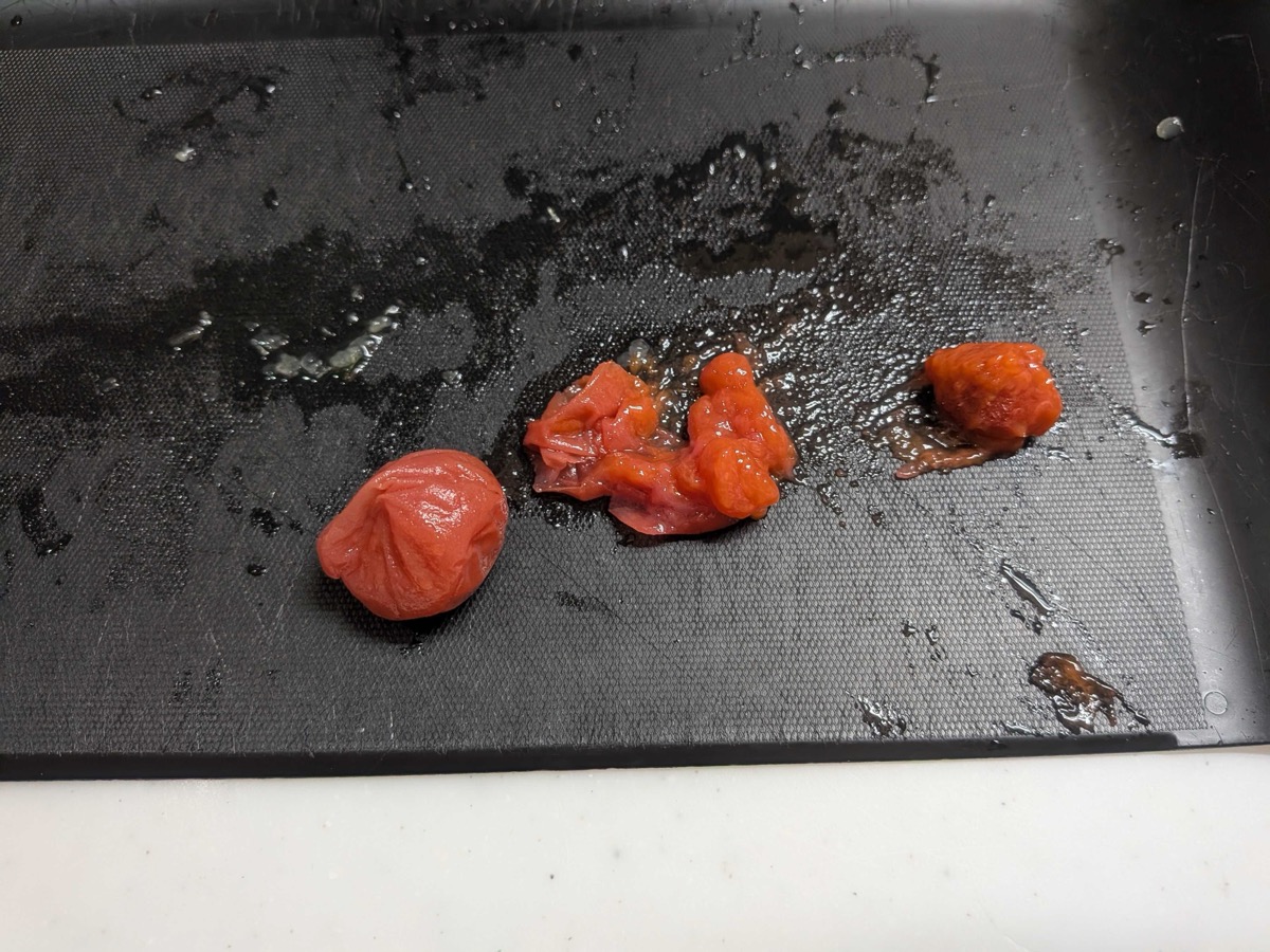 A red umeboshi (salted Japanese plum) roughly chopped into a coarse paste on a black cutting board, next to the whole intact plum