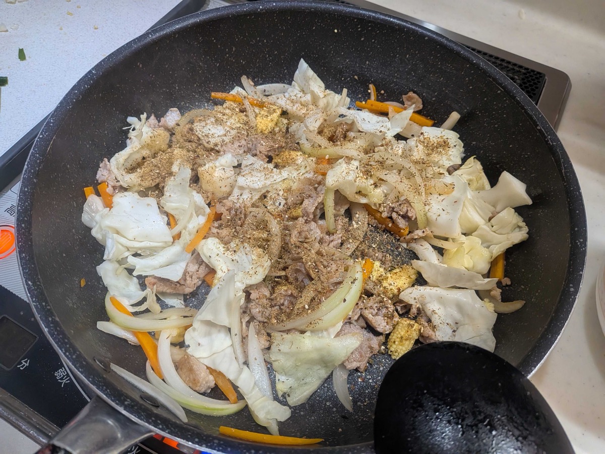 Dashi powder and pepper being sprinkled over stir-fried pork, cabbage, onion, and carrot in a frying pan
