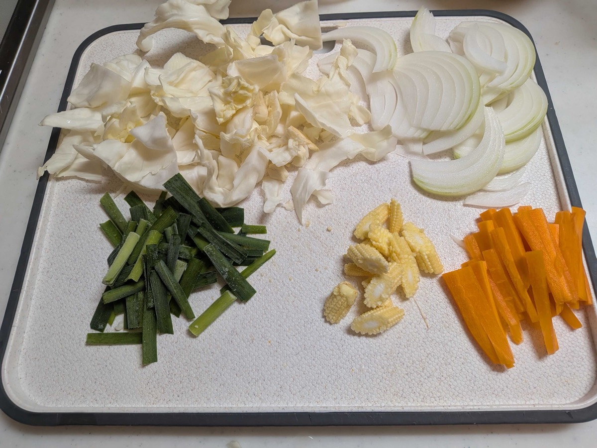 A cutting board with prepped yakiudon vegetables — chopped cabbage, sliced onion, cut garlic chives, halved baby corn, and carrot batons