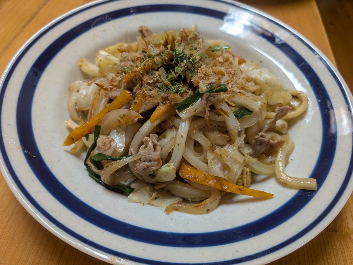 A plate of finished Japanese yakiudon — stir-fried udon noodles with pork, cabbage, carrot, and garlic chives, topped with bonito flakes and green aonori, on a blue-rimmed plate