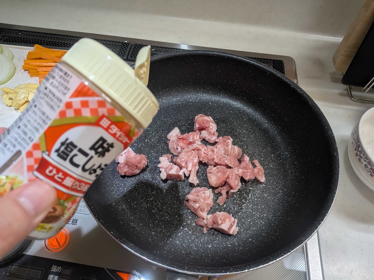 Bite-sized pork pieces cooking in a frying pan, being seasoned with salt and pepper from a shaker