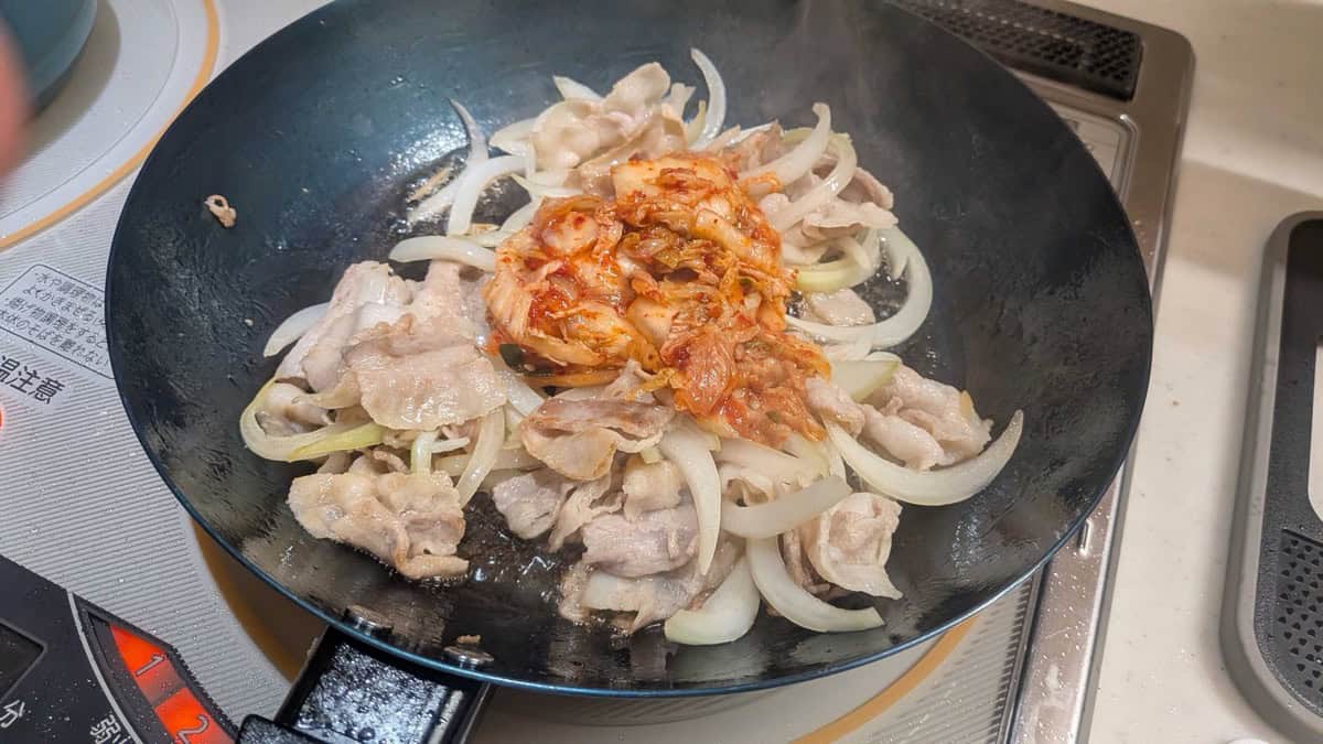 Napa cabbage kimchi added on top of cooked pork and onion in a frying pan