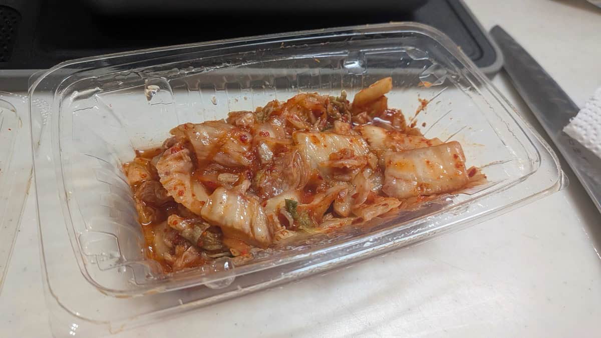 Napa cabbage kimchi in a clear plastic supermarket deli container on a kitchen counter