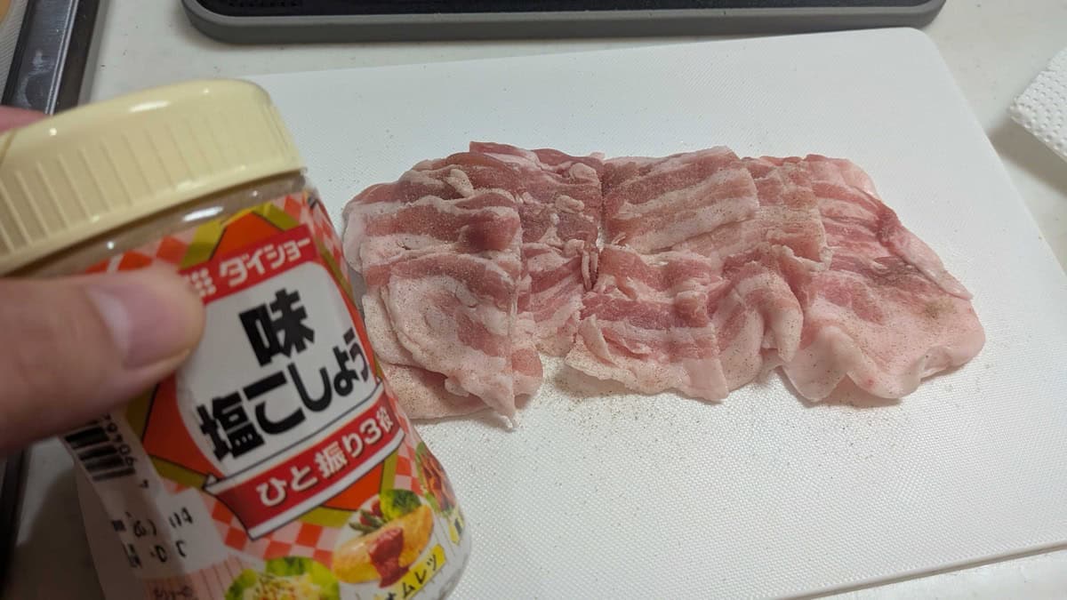 Thin slices of pork belly on a white cutting board, seasoned with Japanese seasoned salt and pepper from a shaker