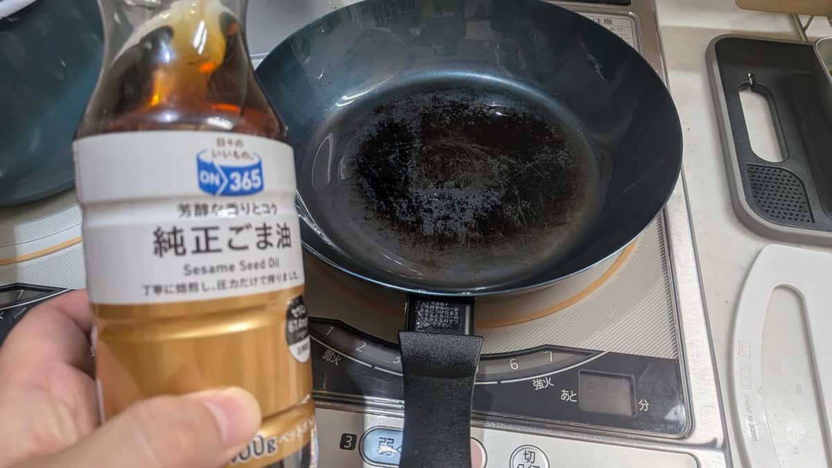 A bottle of pure Japanese sesame oil held next to an empty black frying pan heating on an IH cooktop