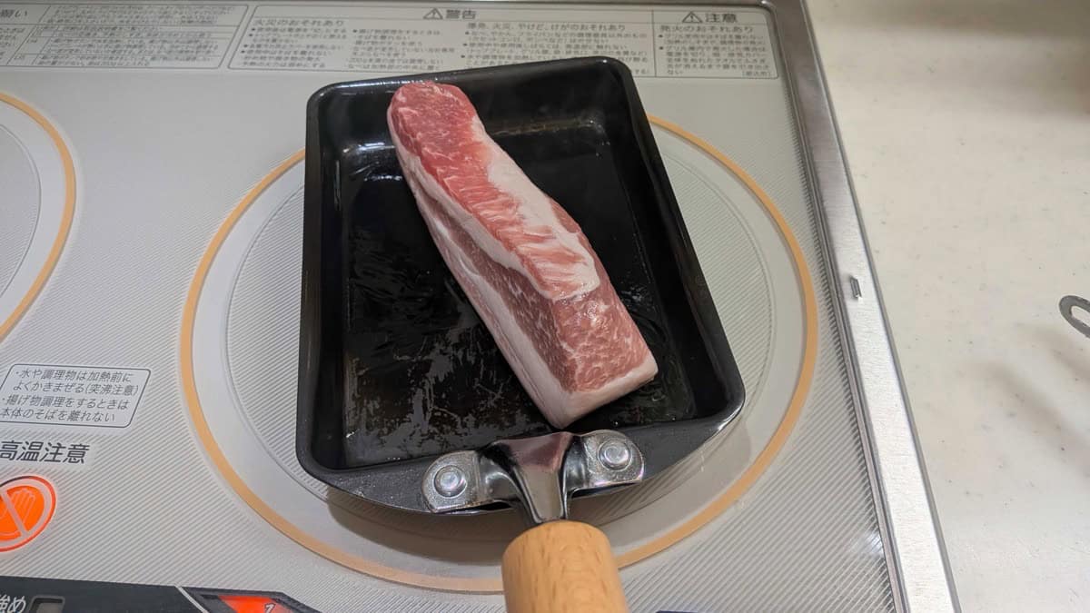 A raw block of pork belly in a small pan on an induction cooktop, ready to sear