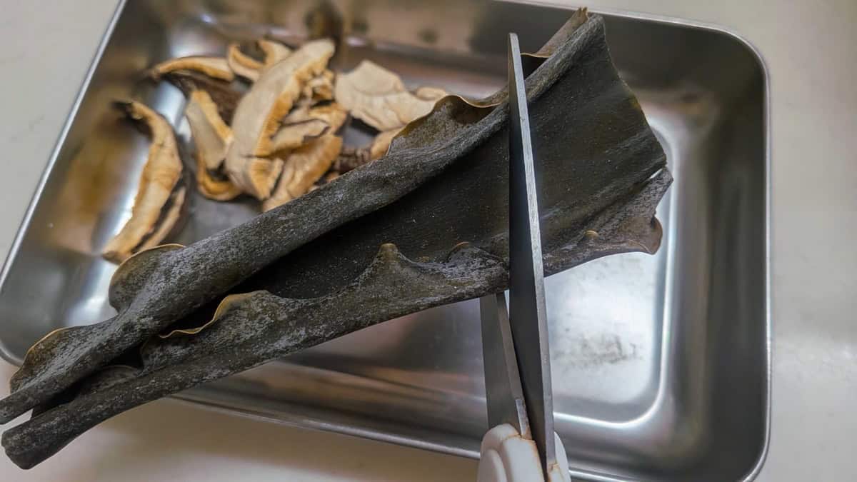 Cutting a strip of dried dashi kombu with kitchen scissors on a metal tray, dried shiitake alongside