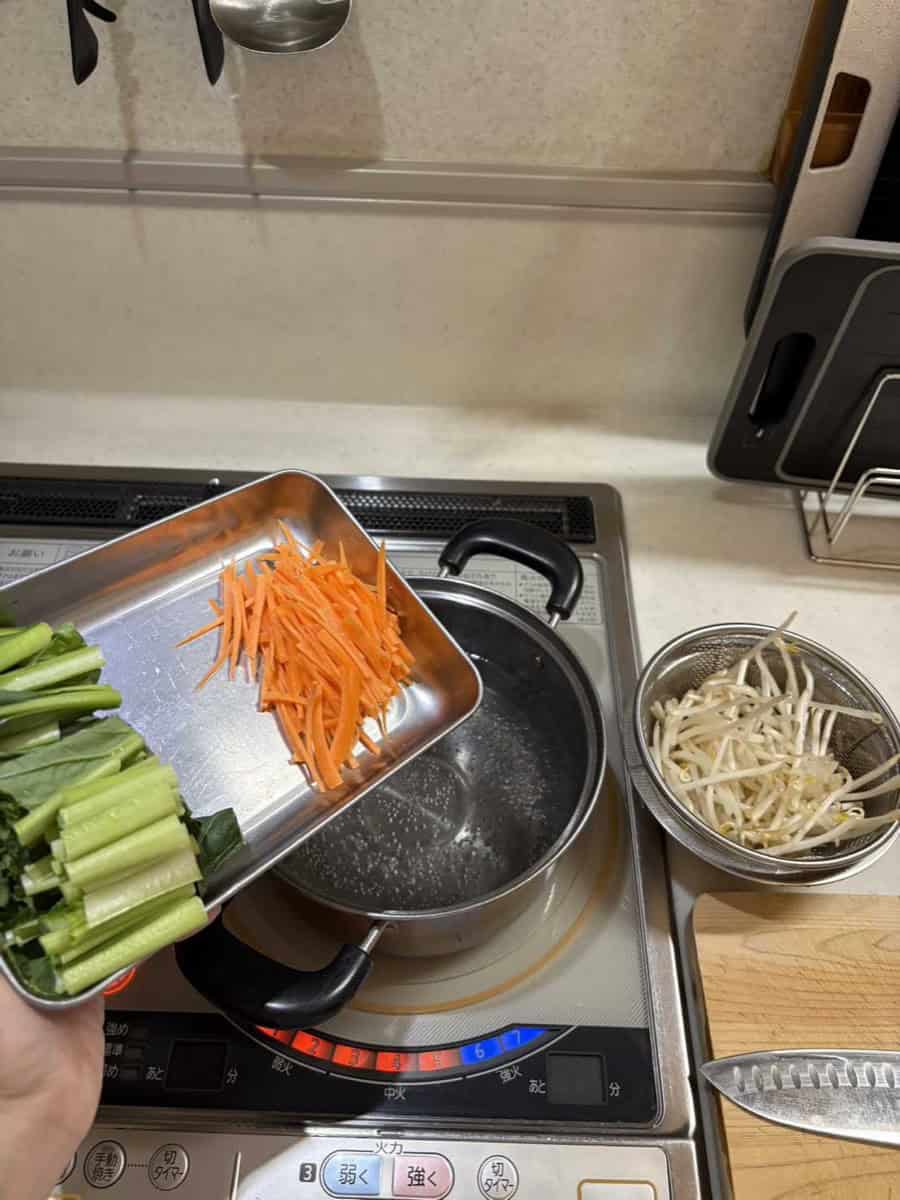 Adding cut komatsuna and carrot to a pot of boiling water, bean sprouts waiting in a strainer