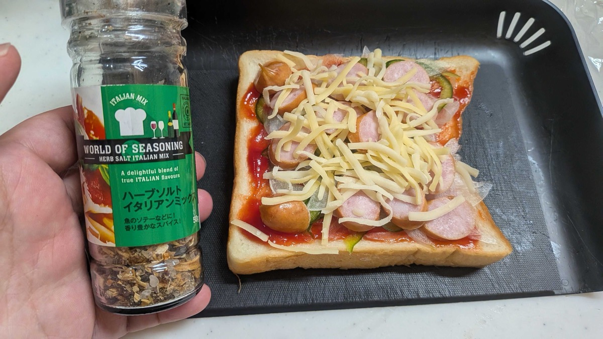 Pizza toast piled with shredded cheese, a bottle of Italian herb salt seasoning held beside it