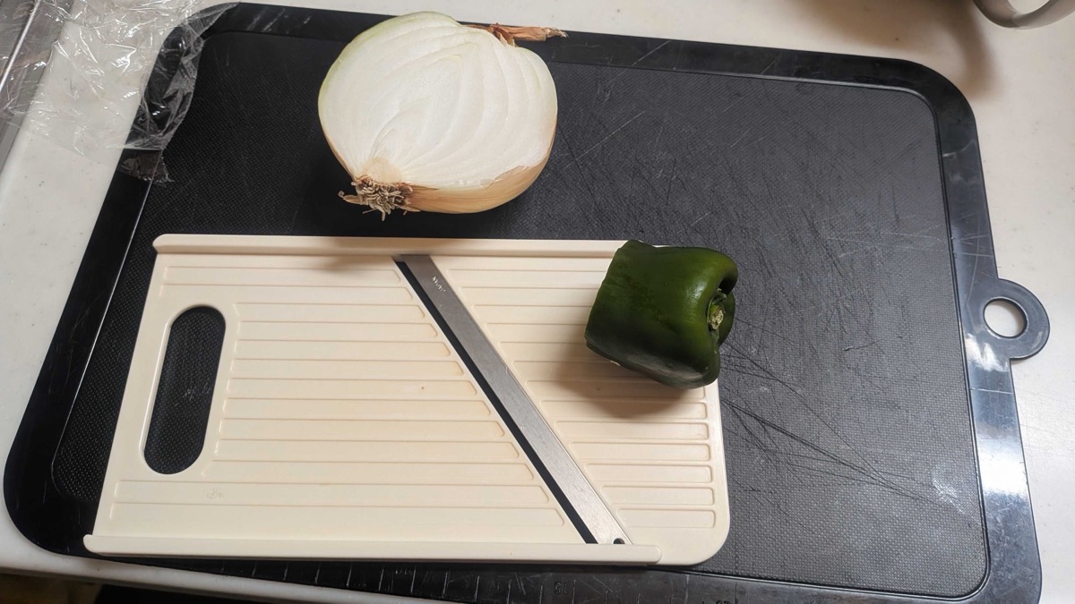 Half an onion and a green pepper next to a Japanese mandoline slicer on a cutting board
