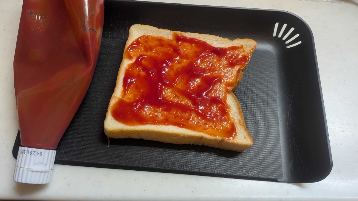 A slice of thick white bread spread with ketchup on a black toaster tray, ketchup bottle beside it