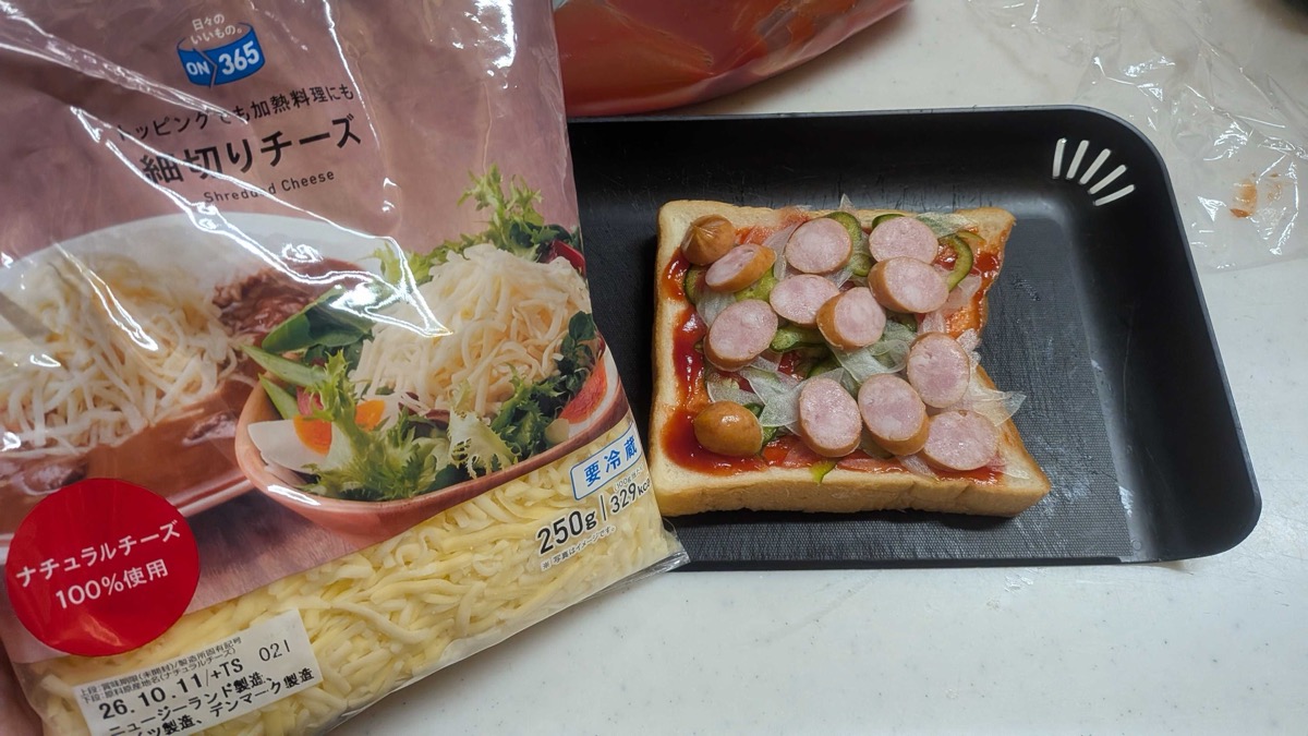 Bread topped with ketchup, thin onion and green pepper, and sliced wiener sausage, next to a bag of shredded cheese