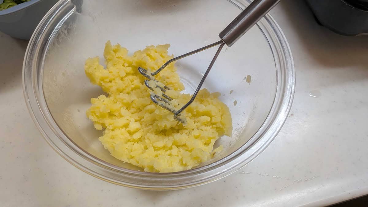 Boiled potatoes partly mashed in a glass bowl with a masher