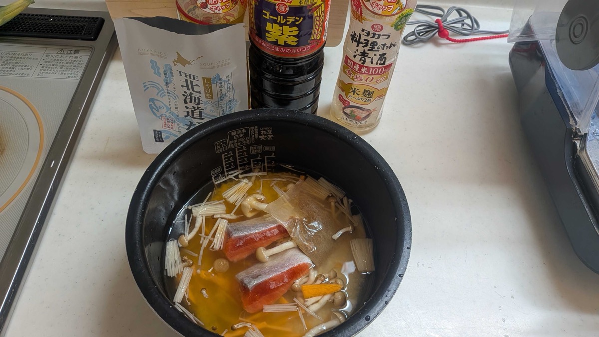 Salmon fillets, enoki and shimeji mushrooms, carrot, and a dashi packet arranged on rice and broth in the rice cooker pot