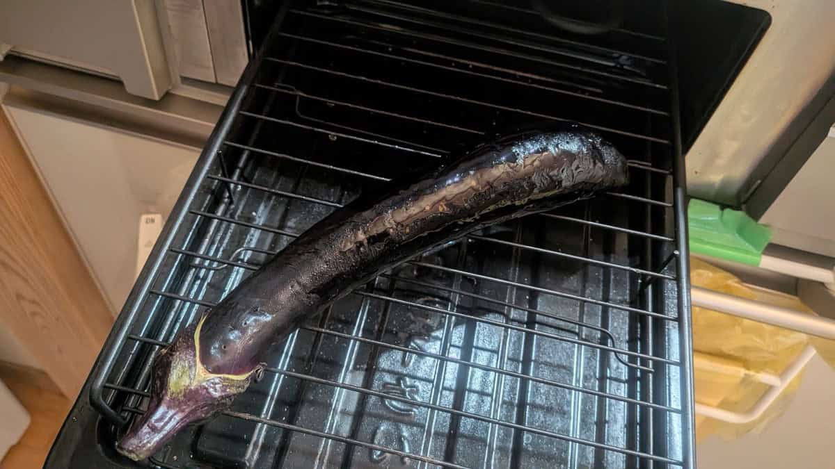 A whole Japanese eggplant blistering on the rack of a fish grill, skin darkened and glossy