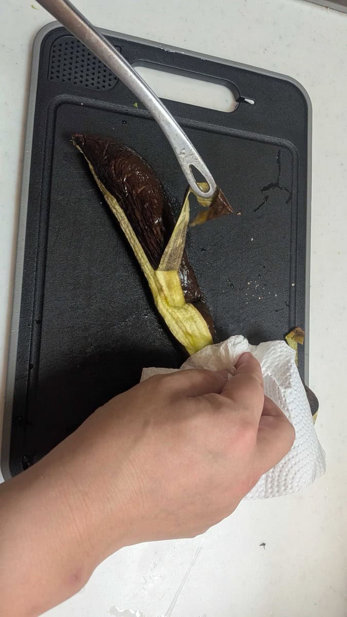Peeling the charred skin off a hot grilled eggplant with a knife, holding the stem with a paper towel