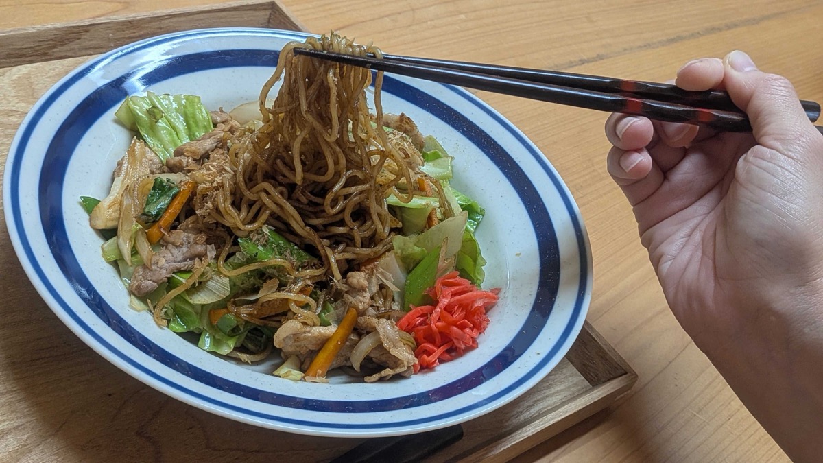 Yakisoba served on a blue-and-white plate with bonito flakes and red pickled ginger, chopsticks lifting the noodles