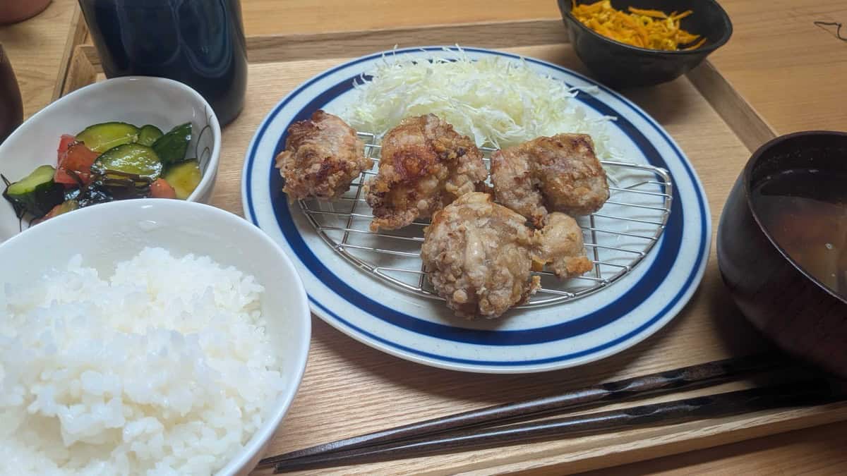 A Japanese home dinner on a wooden tray: air fryer karaage with shredded cabbage, a bowl of rice, miso soup, cucumber and tomato sunomono, and a carrot side dish