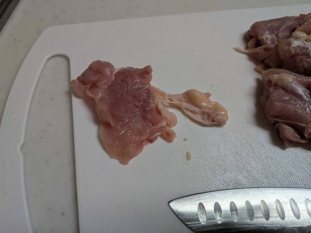Trimming excess fat and sinew from a raw chicken thigh on a white cutting board