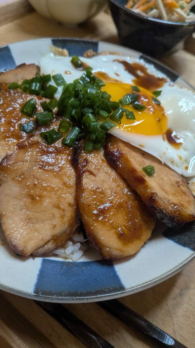 Close-up of glazed chashu slices and a runny fried egg yolk over rice in a blue and white bowl