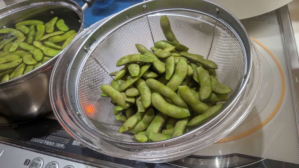 Boiled edamame draining in a metal strainer next to the pot