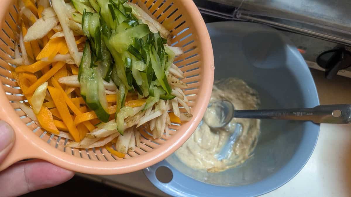 Boiled burdock and carrot with cucumber being tipped from a colander into a bowl of mayonnaise and soy sauce dressing