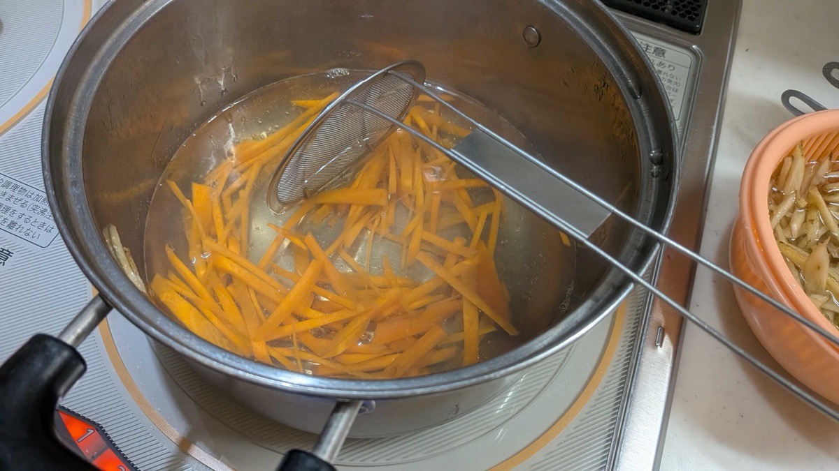 Carrot matchsticks boiling in the same pot with a mesh strainer, with drained burdock in a colander beside the stove