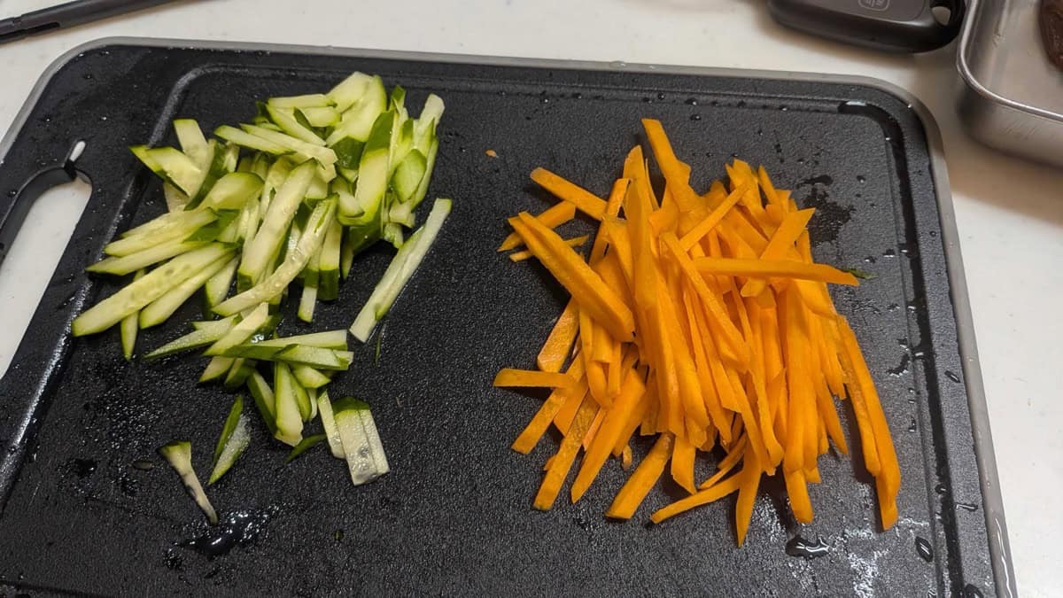 Julienned cucumber and carrot matchsticks side by side on a black cutting board