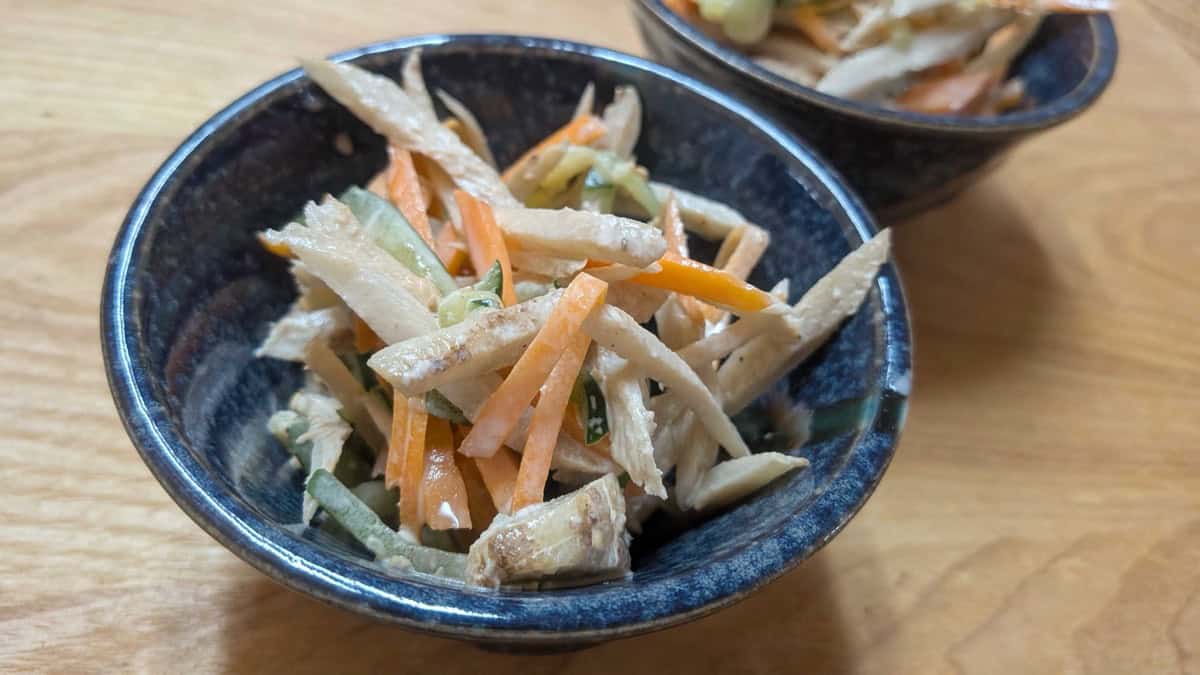 Close view of homemade gobo salad in a small navy bowl on a wooden table, showing burdock, carrot and cucumber strips