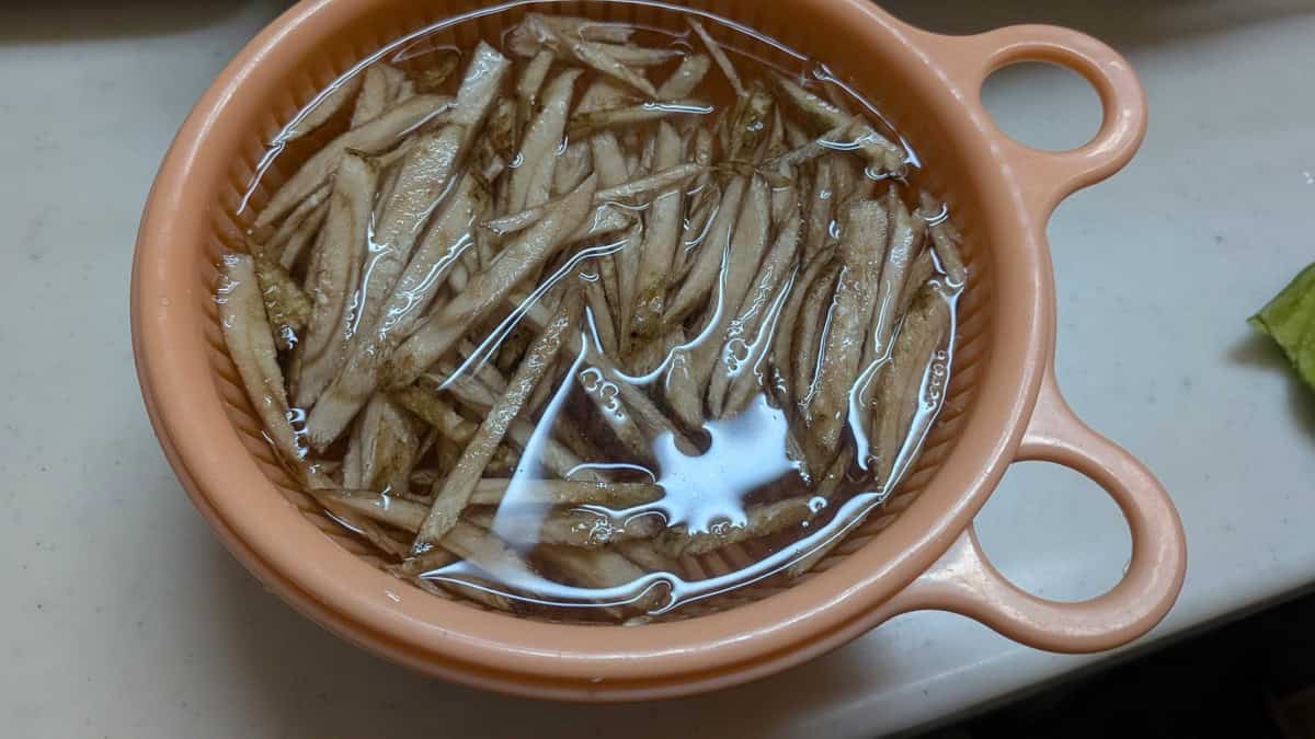 Burdock root matchsticks soaking in water in a pink colander set inside a bowl